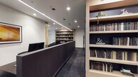 This single-floor space is designed with a contemporary, professional aesthetic that blends functionality with refined style. The interior is anchored by a sleek, dark reception counter that runs along the central corridor, framed by warm natural wood-toned built-in bookshelves stocked with an extensive collection of books. Linear LED pendant lighting and recessed ceiling fixtures illuminate the space consistently throughout, creating a polished and well-lit environment.

A dedicated lounge area features a pair of curved black accent chairs arranged around a round white coffee table, positioned alongside a window that introduces natural light. Built-in shelving units line multiple walls, with dark back panels providing contrast against the light wood framing, and decorative elements such as trailing plants and typographic accents adding character.

A workstation area contains a row of individual computer desks with mesh ergonomic chairs, separated by low partitions against a matte black feature wall, suited for focused individual work. Additional desk space with monitor setups and ergonomic seating is available throughout. Deep charcoal carpet flooring, white walls, and consistent dark furnishings unify the space with a cohesive, modern palette.