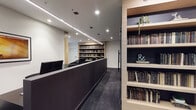 This single-floor space is designed with a contemporary, professional aesthetic that blends functionality with refined style. The interior is anchored by a sleek, dark reception counter that runs along the central corridor, framed by warm natural wood-toned built-in bookshelves stocked with an extensive collection of books. Linear LED pendant lighting and recessed ceiling fixtures illuminate the space consistently throughout, creating a polished and well-lit environment.

A dedicated lounge area features a pair of curved black accent chairs arranged around a round white coffee table, positioned alongside a window that introduces natural light. Built-in shelving units line multiple walls, with dark back panels providing contrast against the light wood framing, and decorative elements such as trailing plants and typographic accents adding character.

A workstation area contains a row of individual computer desks with mesh ergonomic chairs, separated by low partitions against a matte black feature wall, suited for focused individual work. Additional desk space with monitor setups and ergonomic seating is available throughout. Deep charcoal carpet flooring, white walls, and consistent dark furnishings unify the space with a cohesive, modern palette.