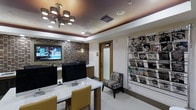 This single-floor space combines functionality with contemporary design. The room is anchored by a sleek white workstation desk equipped with multiple computer monitors, complemented by mustard-yellow upholstered chairs that add a warm pop of color. A large flat-screen television is mounted within a striking mosaic-tiled accent wall featuring earthy brown and beige tones, serving as a bold focal point. An elegant multi-armed chandelier with cylindrical shades hangs from a tray ceiling, which features recessed lighting that enhances the warm, inviting ambiance. A floor-to-ceiling magazine rack along one wall displays an extensive collection of publications, suggesting a waiting or reception area purpose. A cozy patterned armchair is positioned near the center of the room, offering a comfortable seating option. A frosted-glass interior door and a solid wood-framed door provide access to adjoining spaces, adding architectural detail. The light-toned tile flooring ties the room together, contributing to the polished, professional atmosphere throughout the space.