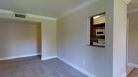 This single-floor residence offers a thoughtfully arranged layout with a warm, neutral color palette throughout. The main living area is finished with plush wall-to-wall carpeting and features elegant crown molding that adds a refined architectural detail to the space. Clean white baseboards frame the room, providing a crisp contrast against the soft, beige-toned walls.

A standout feature of the living area is the kitchen pass-through window, which creates an open visual connection between the two spaces. The kitchen is equipped with dark wood cabinetry, a white over-the-range microwave, and a white electric stove, all accented by a stone-topped countertop ledge at the pass-through. The textured ceiling adds subtle visual depth to the interior.

An open doorway leads from the main living area into an adjoining room, suggesting a versatile and connected floor plan that allows for natural flow between spaces. The overall ambiance is clean, understated, and functional, making it a practical and comfortable single-floor living environment.