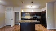 Located at 3245 Glen McConnell Parkway in Charleston, South Carolina, this single-floor residence offers a thoughtfully designed layout that balances functionality and style. The kitchen serves as a central highlight of the space, showcasing rich dark cabinetry that contrasts beautifully against light-toned walls. A substantial center island with a granite-style countertop provides generous prep space and doubles as a casual gathering spot. Pendant lighting hangs above the island, adding warm, focused illumination that enhances the kitchen's inviting atmosphere. A built-in wine rack adds a distinctive decorative touch to the cabinetry. Full black appliances, including a refrigerator, range, and microwave, are seamlessly integrated into the kitchen design. Warm hardwood-style flooring flows throughout, lending continuity and a natural warmth to the interior. A walk-in pantry or utility closet is conveniently accessible just off the kitchen, offering practical storage solutions. The overall ambiance of the space is warm and welcoming, with a clean, contemporary aesthetic that makes it well-suited for both everyday living and entertaining.