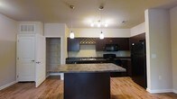 Located at 3245 Glen McConnell Parkway in Charleston, South Carolina, this single-floor residence offers a thoughtfully designed layout that balances functionality and style. The kitchen serves as a central highlight of the space, showcasing rich dark cabinetry that contrasts beautifully against light-toned walls. A substantial center island with a granite-style countertop provides generous prep space and doubles as a casual gathering spot. Pendant lighting hangs above the island, adding warm, focused illumination that enhances the kitchen's inviting atmosphere. A built-in wine rack adds a distinctive decorative touch to the cabinetry. Full black appliances, including a refrigerator, range, and microwave, are seamlessly integrated into the kitchen design. Warm hardwood-style flooring flows throughout, lending continuity and a natural warmth to the interior. A walk-in pantry or utility closet is conveniently accessible just off the kitchen, offering practical storage solutions. The overall ambiance of the space is warm and welcoming, with a clean, contemporary aesthetic that makes it well-suited for both everyday living and entertaining.