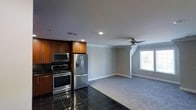 This single-floor space offers an open-concept layout that seamlessly connects the kitchen and living area, creating a functional and cohesive environment. The kitchen is well-appointed with stainless steel appliances, including a gas range with oven, an over-the-range microwave, and a French door refrigerator with an ice and water dispenser. Rich, warm-toned wood cabinetry provides ample storage and adds a touch of traditional character to the space. The kitchen floor is finished with large, dark polished tiles that contrast elegantly with the lighter tones throughout the rest of the space. Transitioning into the living area, the flooring shifts to a light-toned carpet, contributing to a comfortable and welcoming atmosphere. Crown molding runs along the ceiling, adding a refined architectural detail to the room. A ceiling fan with a light fixture is centrally positioned in the living area, offering both air circulation and ambient lighting. A window allows natural light to fill the space, enhancing the airy feel of the open layout. Recessed lighting in the kitchen provides focused illumination, ensuring the space is both practical and well-lit throughout.