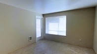 This single-story space features a straightforward, functional layout with a clean and neutral aesthetic throughout. The main living area is finished with wall-to-wall carpet in a light beige tone, contributing to a quiet and understated atmosphere. Walls are painted in a soft gray-beige, providing a versatile backdrop suitable for a variety of furnishing styles.

Natural light is a notable feature of this space, with a large picture window fitted with horizontal blinds allowing for adjustable brightness. An exterior door adjacent to the window also includes a blind-covered glass panel, further enhancing the flow of natural light into the room while providing direct outdoor access.

A floor-level heating vent is positioned along the base of one wall, indicating a forced-air heating system integrated into the unit. The ceilings are smooth and white, adding to the overall sense of openness in the room.

Overall, the space presents a simple, move-in-ready environment with practical finishes and adequate natural light, well-suited for comfortable everyday living.