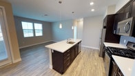 This single-floor space showcases a thoughtfully designed open-concept layout that seamlessly connects the kitchen and living areas. The kitchen serves as a central focal point, anchored by a substantial island with a crisp white countertop, an undermount sink, and dark espresso-toned cabinetry with brushed metal hardware. Two sleek pendant lights hang above the island, providing both functional task lighting and a modern aesthetic touch. Stainless steel appliances complement the contemporary design palette.

The flooring throughout is a warm, light wood-style material that adds continuity and brightness across the open plan. Recessed lighting in the ceiling supplements the natural light that filters in through windows along the far wall of the living area, creating a well-lit and airy atmosphere. Neutral gray walls provide a clean, versatile backdrop that enhances the sense of openness.

The living area flows naturally from the kitchen, offering generous space for furniture arrangement. The overall design is clean and modern, characterized by consistent finishes, a neutral color scheme, and an efficient use of space that makes the interior feel both functional and inviting.