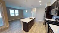 This single-floor space showcases a thoughtfully designed open-concept layout that seamlessly connects the kitchen and living areas. The kitchen serves as a central focal point, anchored by a substantial island with a crisp white countertop, an undermount sink, and dark espresso-toned cabinetry with brushed metal hardware. Two sleek pendant lights hang above the island, providing both functional task lighting and a modern aesthetic touch. Stainless steel appliances complement the contemporary design palette.

The flooring throughout is a warm, light wood-style material that adds continuity and brightness across the open plan. Recessed lighting in the ceiling supplements the natural light that filters in through windows along the far wall of the living area, creating a well-lit and airy atmosphere. Neutral gray walls provide a clean, versatile backdrop that enhances the sense of openness.

The living area flows naturally from the kitchen, offering generous space for furniture arrangement. The overall design is clean and modern, characterized by consistent finishes, a neutral color scheme, and an efficient use of space that makes the interior feel both functional and inviting.