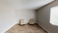 This single-floor space presents a straightforward and functional layout with a clean, neutral aesthetic throughout. The main living area features warm-toned hardwood-style flooring that runs seamlessly across the room, providing a grounded and inviting foundation. The walls are finished in a soft, neutral gray-beige tone that lends a calm and versatile backdrop, easily adaptable to a variety of furnishing styles and décor preferences. A classic textured popcorn ceiling adds a familiar residential character to the space. Crisp white baseboards provide a clean contrast against both the walls and flooring, contributing to the room's tidy, well-maintained appearance. The room is generously proportioned, offering ample open floor space with room to accommodate a variety of furniture arrangements. The overall ambiance is understated and move-in ready, with a simple and practical design that prioritizes flexibility. Whether used as a living area, sitting room, or multipurpose space, the room's neutral palette and durable finishes make it a solid and adaptable environment suited to a range of uses.