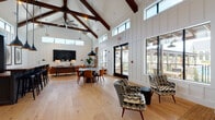 This single-story space is defined by its dramatic vaulted ceiling supported by richly stained exposed wood trusses, giving the interior a barn-inspired architectural character. White shiplap and board-and-batten wall paneling run throughout, creating a clean, modern farmhouse aesthetic that contrasts beautifully with the warm-toned wide-plank hardwood floors underfoot.

The open floor plan accommodates several distinct functional zones. A central bar area serves as an anchor point, featuring a dark cabinetry surround, a rich wood countertop, bar-height seating, and a decorative blue-and-white patterned tile backsplash illuminated by industrial pendant lights. Adjacent to the bar, a dining area provides a round table with amber upholstered chairs, while a lounge zone offers leather seating, a large-format wall-mounted television, and curated artwork.

A quieter seating nook near the perimeter windows features bold patterned accent chairs alongside a built-in bench, providing an intimate retreat within the larger space. Clerestory windows positioned high along the walls, combined with large black-framed windows and glass doors, flood the interior with abundant natural light and offer outdoor views of a surrounding covered patio area.