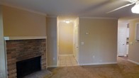 This single-story space combines classic design elements with practical functionality. The living area is anchored by a traditional brick fireplace with a clean white mantle surround, serving as a focal point within the room. Neutral beige walls paired with white crown molding and white baseboards lend the space a timeless, cohesive aesthetic. The living area features wall-to-wall carpeting in a warm tan tone, contributing to a cozy atmosphere.

A hallway extends from the main living space, transitioning to tile flooring and connecting to additional areas of the home. The hallway is lit by a flush-mount ceiling light, providing consistent illumination throughout. Doors along the hallway suggest access to closet storage and additional rooms, enhancing the overall functionality of the layout.

Throughout the space, crown molding adds a refined architectural detail, while the neutral color palette creates a versatile backdrop. The single-floor layout ensures seamless flow between areas, making the space practical and easy to navigate. Overall, the design balances understated elegance with everyday comfort.