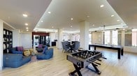 This single-floor communal space is thoughtfully designed to accommodate a variety of activities, blending recreational and social zones into one open, well-appointed layout. The interior features light hardwood-style flooring throughout, recessed ceiling lighting, and a tray ceiling detail that adds architectural dimension while defining different areas within the space.

At the forefront of the room, a foosball table serves as a central recreational focal point, complemented by a pool table visible toward the rear, making this area well-suited for group entertainment. Adjacent to the game area, a casual lounge zone features plush, low-profile seating with decorative accent pillows arranged over a patterned area rug, offering a comfortable setting for informal gatherings.

A dining area with upholstered chairs around a round table provides a more structured seating option, while a vending machine station and a coffee or refreshment counter along the back wall ensure convenient access to beverages and snacks. Large windows along one side of the room allow natural light to filter in, contributing to the bright and welcoming ambiance of the overall space.