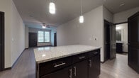 This single-floor unit offers a thoughtfully designed layout with modern finishes throughout. The kitchen serves as a central highlight, featuring dark espresso cabinetry with brushed nickel hardware, granite countertops, and a full suite of stainless steel appliances including a range, over-the-range microwave, dishwasher, and refrigerator. A substantial kitchen island with matching granite provides additional prep and seating space, while dual pendant lights overhead add focused illumination. A decorative wine rack accent adds a distinctive touch to the upper cabinetry. Light wood-style flooring flows through the kitchen and into the open living area, which is equipped with a ceiling fan and offers natural light through blinded windows. The bedrooms feature plush carpeting, ceiling fans, and generous window arrangements that invite natural light while maintaining privacy. Dark-trimmed doors provide a consistent design element throughout the unit. The bathroom continues the cohesive aesthetic with dark cabinetry, granite countertops, a large mirror, and well-placed vanity lighting. A programmable thermostat is conveniently located in the living area. Overall, the unit presents a clean, contemporary feel with a neutral color palette and a functional, open flow.