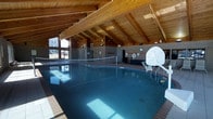 This indoor recreational facility centers around a generously sized swimming pool housed beneath a striking vaulted ceiling constructed with exposed wood beams and tongue-and-groove paneling, lending the space a warm, lodge-like ambiance. The soaring roofline creates an open, airy atmosphere that prevents the enclosed pool area from feeling confined. The pool itself is equipped for active recreation, featuring a water volleyball net stretched across its width and a poolside basketball hoop, making it well-suited for both leisure and organized aquatic activities. Tiled walls line the perimeter of the space, providing a durable, easy-to-maintain surface that complements the natural wood overhead. Safety equipment, including life rings, is visibly mounted on the walls, and a clock keeps swimmers and guests informed of the time. Large windows at one end of the facility allow natural light to filter in, brightening the water and enhancing the overall atmosphere. The deck surrounding the pool provides ample space for lounging or moving between activities. This versatile indoor aquatic center is designed to accommodate a wide range of recreational uses year-round, regardless of outdoor weather conditions.