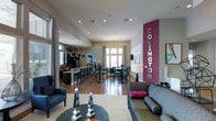 The Covington clubhouse is a thoughtfully designed single-floor amenity space that seamlessly blends social and functional areas into one cohesive environment. A bold magenta accent column bearing the "Covington" name serves as a striking focal point, immediately establishing a strong sense of place and identity upon entry.

The lounge area features a patterned sofa with accent pillows, a navy blue armchair, and a round coffee table dressed with decorative obelisk sculptures atop a neutral area rug, creating a comfortable and stylish gathering spot. Warm hardwood flooring extends throughout, unifying the space visually.

Adjacent to the lounge, a dining area accommodates multiple guests with dark wood high-top tables and chairs arranged beneath a classic chandelier, with large windows allowing natural light to fill the room. Neutral drapes frame the windows, softening the overall aesthetic.

The kitchen bar area runs along one side of the space, equipped with pendant lighting, bar-height seating, and stainless steel appliances, making it well-suited for catered events or community gatherings. Recessed lighting throughout ensures consistent illumination across all zones, contributing to the space's open and inviting ambiance.