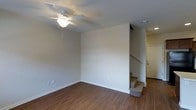 This two-story space offers a straightforward and functional layout designed for comfortable everyday living. The main level features warm hardwood-style flooring that extends throughout the living area, complementing the clean, neutral-toned walls that create a bright and open atmosphere. A ceiling fan with a light fixture is centrally mounted, providing both airflow and ambient lighting to the room.

A carpeted staircase is positioned toward the back of the main level, providing access to the upper floor. The staircase is framed by a partial wall, adding a sense of structure and definition to the living space. White baseboards run along the perimeter of the room, offering a clean contrast to the flooring.

Electrical outlets are conveniently placed along the walls, and the overall design maintains a simple, minimalist aesthetic with neutral paint throughout. A doorway visible near the staircase suggests additional rooms or storage on the main level. The upper floor, accessible via the staircase, expands the functional living space of this two-story layout.