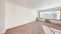 This single-floor studio unit offers a practical and functional open-concept layout that combines the living and kitchen areas into one cohesive space. The main living area is generously sized, featuring wall-to-wall carpet in a neutral tone and crisp white walls that create a clean, bright atmosphere. A large picture window with a warm wood frame anchors one wall, flooding the room with abundant natural light and providing views of the outdoors. A wall-mounted HVAC unit and baseboard heating ensure year-round climate comfort.

The kitchen is neatly integrated into an alcove at one end of the main room, defined by warm wood cabinetry above and below. It is equipped with a white gas range, a full-size refrigerator, and a single-basin sink with a wood-toned cabinet base, providing all essential cooking and storage needs in a compact footprint.

A louvered bifold closet door near the entryway offers convenient storage. A hallway connects the main space to the bathroom, which features a full-size soaking tub surrounded by clean white square tile, a grab bar, and a simple, well-maintained design. The unit's straightforward layout and abundant natural light make it a highly functional and comfortable living space.