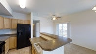 Located at 7702 Little Fox Ln in Wellington, CO, this single-floor home offers a thoughtfully designed open-concept layout that seamlessly connects the kitchen and living areas. The kitchen is equipped with light wood cabinetry, a black refrigerator, and a wraparound peninsula countertop featuring a built-in sink — ideal for both meal preparation and casual dining. The countertops carry a warm, earthy tone that complements the tile flooring in the kitchen area. The living space transitions naturally from the kitchen and is finished with neutral carpet flooring, creating a comfortable and cohesive feel. A ceiling fan with an integrated light fixture is centrally positioned overhead, providing both air circulation and ambient lighting. Large sliding glass doors flood the living area with natural light and open directly onto a balcony, offering outdoor views and a seamless indoor-outdoor connection. A hallway visible from the main living area leads to additional spaces within the home, including what appears to be a laundry or storage area with a white door. The overall ambiance of the home is bright, clean, and functional, with a neutral color palette that lends itself to a variety of interior styles.