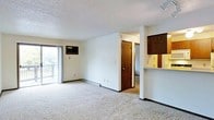 This single-floor unit offers a practical, open layout centered around a generously sized living area finished with light-colored wall-to-wall carpeting. The textured ceiling and neutral white walls provide a clean, simple backdrop throughout the space. A sliding glass door along one wall floods the living area with natural light and opens to an outdoor balcony, offering views of surrounding greenery. A wall-mounted air conditioning unit is positioned nearby for convenient climate control, accompanied by a baseboard heating system along the perimeter walls.

A half-wall partition separates the living area from the kitchen, featuring a pass-through counter that enhances the open feel between the two spaces. The kitchen is equipped with dark wood cabinetry, providing ample storage and a warm contrast to the light surroundings. A doorway visible from the living area leads further into the unit, connecting to additional rooms beyond. The overall layout is efficient and straightforward, making it easy to navigate between spaces. The unit's design prioritizes functionality, with clean lines, ample natural light, and practical finishes throughout.