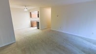 This single-floor space features an open-concept layout that seamlessly connects the living area to the kitchen. The living area is generously sized, with soft, neutral-toned wall-to-wall carpeting that adds warmth underfoot. White walls and clean baseboards create a bright, understated backdrop throughout the space. A ceiling fan is installed overhead, providing both lighting and air circulation.

The kitchen is partially open to the living area, divided by a half-wall that maintains a sense of connection between the two spaces. It features warm wood-toned cabinetry offering ample storage, a countertop workspace, and a stainless steel appliance — likely a dishwasher — visible along the lower cabinet line. The kitchen area transitions to a hard flooring surface, contrasting with the carpeted living space.

Overall, the space presents a functional and straightforward layout with a neutral color palette. The open sightlines between the kitchen and living area contribute to an airy feel, making the space feel cohesive and practical for everyday living.