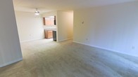 This single-floor space features an open-concept layout that seamlessly connects the living area to the kitchen. The living area is generously sized, with soft, neutral-toned wall-to-wall carpeting that adds warmth underfoot. White walls and clean baseboards create a bright, understated backdrop throughout the space. A ceiling fan is installed overhead, providing both lighting and air circulation.

The kitchen is partially open to the living area, divided by a half-wall that maintains a sense of connection between the two spaces. It features warm wood-toned cabinetry offering ample storage, a countertop workspace, and a stainless steel appliance — likely a dishwasher — visible along the lower cabinet line. The kitchen area transitions to a hard flooring surface, contrasting with the carpeted living space.

Overall, the space presents a functional and straightforward layout with a neutral color palette. The open sightlines between the kitchen and living area contribute to an airy feel, making the space feel cohesive and practical for everyday living.