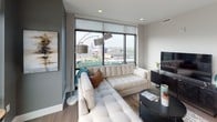 Located at 338–398 Bridge St NW in Grand Rapids, Michigan, this single-floor unit is situated at an elevated position within its building, offering striking panoramic views of the Grand Rapids skyline and surrounding urban landscape. The living area is anchored by expansive floor-to-ceiling windows fitted with roller shades, flooding the space with natural light while framing broad city views. Wood-look flooring, neutral gray walls, and recessed lighting contribute to a clean, modern aesthetic throughout.

The kitchen is designed with functionality and style in mind, featuring flat-front white cabinetry extending to the ceiling, white quartz countertops, a gray tile backsplash, stainless steel appliances, and a window positioned above the sink. The layout provides generous counter space and ample storage.

Multiple bedrooms are present, each finished with wood-look flooring and neutral tones. The primary bedroom includes a sliding barn-style door opening to a walk-in closet and windows offering city views. A secondary bedroom features a sliding frosted-panel closet door and its own window with green treetop views.

The bathroom includes a tiled soaking tub and shower combination, wood-look flooring, a light-framed vanity mirror, and a white quartz-topped vanity, maintaining the unit's cohesive modern character.