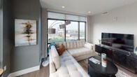 Located at 338–398 Bridge St NW in Grand Rapids, Michigan, this single-floor unit is situated at an elevated position within its building, offering striking panoramic views of the Grand Rapids skyline and surrounding urban landscape. The living area is anchored by expansive floor-to-ceiling windows fitted with roller shades, flooding the space with natural light while framing broad city views. Wood-look flooring, neutral gray walls, and recessed lighting contribute to a clean, modern aesthetic throughout.

The kitchen is designed with functionality and style in mind, featuring flat-front white cabinetry extending to the ceiling, white quartz countertops, a gray tile backsplash, stainless steel appliances, and a window positioned above the sink. The layout provides generous counter space and ample storage.

Multiple bedrooms are present, each finished with wood-look flooring and neutral tones. The primary bedroom includes a sliding barn-style door opening to a walk-in closet and windows offering city views. A secondary bedroom features a sliding frosted-panel closet door and its own window with green treetop views.

The bathroom includes a tiled soaking tub and shower combination, wood-look flooring, a light-framed vanity mirror, and a white quartz-topped vanity, maintaining the unit's cohesive modern character.