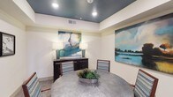 This single-story space functions as a well-appointed meeting or private dining room, thoughtfully designed with a cohesive aesthetic that balances professionalism with warmth. The room's most striking architectural feature is its tray ceiling, finished in a deep teal-blue tone that contrasts sharply against the crisp white upper walls, adding a sense of depth and visual interest overhead. Recessed lighting is embedded within the tray, providing focused, even illumination throughout the space.

At the center of the room sits a substantial round table with a hammered metallic finish, surrounded by wood-framed upholstered chairs featuring a blue-toned woven fabric. A low tray centerpiece with decorative succulents adds a natural, refined touch to the table setting.

The walls are adorned with carefully selected artwork. Large abstract paintings in soft blues, greens, and whites anchor two walls, evoking a serene, nature-inspired atmosphere. On an adjacent wall, a series of four black-framed clock-face photographs introduces a graphic, monochromatic contrast. A dark wood sideboard with matching teal-accented lamps completes the furnishings.

The room is accessed through glass-paneled double doors with dark frames, allowing views into the adjoining corridor and hinting at a broader facility beyond.