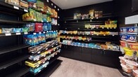 This single-floor convenience market is designed with a sleek, modern aesthetic, defined by its striking matte black walls, shelving units, and cabinetry throughout. The compact yet well-organized layout maximizes every inch of available space, creating distinct product zones that guide shoppers efficiently through the store.

The snack and candy section dominates one wall, with floor-to-ceiling black slatwall and shelving displaying a broad assortment of chips, cookies, candy bars, bagged candy, and protein and nutrition bars. Lower cabinets with brushed hardware provide additional storage beneath the display shelves. An adjacent wall hosts pantry staples, canned goods, condiments, laundry detergent, pet supplies, and other household necessities, organized on sturdy pegboard-backed shelving.

A dedicated refrigeration zone lines another wall, featuring multiple glass-door coolers stocked with a wide variety of bottled beverages, including sodas, juices, and water. An additional upright refrigerator unit provides further cold storage for perishable items. Personal care and miscellaneous convenience products are merchandised on pegboard near the entrance.

The store is accessed through a glass-panel door, with light wood-look flooring providing a clean contrast to the dark interior palette. Recessed and fluorescent lighting illuminates the merchandise effectively, creating a bright, inviting shopping environment.