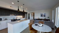 This single-floor space showcases a thoughtfully designed open-concept layout that seamlessly integrates the kitchen, dining, and living areas into one cohesive environment. The kitchen is a standout feature, equipped with dark espresso cabinetry that extends to the ceiling, a light quartz countertop, and a generous island that doubles as a workspace and casual gathering spot. A light-toned tile backsplash complements the cabinetry, while stainless steel appliances add a professional touch. Two striking black pendant lights with gold interiors hang above the island, providing both task lighting and a bold visual accent.

Adjacent to the kitchen, the dining area centers around a round marble-topped table paired with cognac leather chairs, offering an elegant yet inviting spot for meals. The living area beyond is furnished with a plush gray sectional sofa, a wooden media console, and framed artwork, creating a relaxed and curated atmosphere. Recessed lighting throughout maintains a clean ceiling line, while light wood-style flooring runs continuously across the entire space, tying each zone together and contributing to an airy, modern aesthetic.