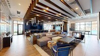 Located at 3600 W Happy Valley Rd in Phoenix, AZ, this single-floor community clubhouse offers a generously sized, open-plan gathering space defined by sophisticated contemporary design. Warm hardwood-style flooring flows continuously throughout, complementing the striking layered wood beam ceiling detail that serves as the room's architectural centerpiece. The main lounge area features an oversized sectional sofa, accent chairs in bold navy blue, and a patterned area rug, creating an inviting social hub. A built-in bar with counter seating and a wall-mounted television anchors one end of the space, while a sleek linear fireplace framed by a concrete surround and decorative wood log storage adds warmth and visual interest to another zone. A full-size billiards table occupies its own dedicated area, bathed in natural light from large windows that frame outdoor pool views. Vertical wood slat partitions elegantly define intimate nooks without closing off the open feel. Throughout the space, curated artwork and sculptural lighting fixtures—including dramatic pendant clusters and linear drops—add artistic flair. The entry corridor leads guests seamlessly into the expansive interior, which benefits from abundant natural light and high ceilings, creating an airy, upscale atmosphere ideal for gathering and entertaining.