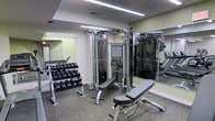 This fitness center is a thoughtfully designed workout facility housed on a single level, offering a comprehensive range of exercise equipment to support a full-body training regimen. The space is laid out to maximize functionality, with distinct zones dedicated to cardio and strength training.

The cardio area features a row of treadmills positioned centrally, alongside additional cardiovascular machines including ellipticals and step machines, providing users with multiple options for aerobic exercise. The flooring throughout the gym is a durable, rubberized surface well-suited to heavy equipment and high-traffic use.

The strength training zone is equally well-appointed, showcasing a multi-station cable machine that supports a wide variety of resistance exercises. Adjustable workout benches are positioned nearby for versatility. A tiered dumbbell rack lines the wall, stocked with a full range of free weights to accommodate all fitness levels.

Full-length mirrors are installed along the walls, enhancing the sense of space while allowing users to monitor their form during workouts. Overhead linear lighting provides bright, even illumination throughout the room. The walls feature a soft neutral tone, contributing to a clean and functional aesthetic. Overall, this fitness center is a well-organized and fully equipped space designed to meet a broad range of fitness needs.