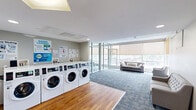Located at 1000 Diamond St in Philadelphia's 19122 neighborhood, this amenity floor offers a thoughtfully designed, multi-functional space spread across a single level. The layout combines three distinct areas that cater to both practical and collaborative needs.

The laundry room is equipped with a row of modern front-loading washers and dryers, set against light walls and finished with warm hardwood flooring. Informational signage and ample counter space make the room both functional and user-friendly.

Adjacent to the laundry area is a bright, open lounge finished with patterned blue carpet tiles. Comfortable grey sofas and armchairs are arranged to take full advantage of the expansive floor-to-ceiling windows, which frame sweeping views of the surrounding Philadelphia cityscape and rail corridor, filling the space with abundant natural light. Roller shades allow for adjustable light control.

A glass-walled conference and study room sits at the far end of the floor, enclosed by sleek aluminum-framed glass partitions. Inside, the room is outfitted with desks, ergonomic task chairs, a wall-mounted flat-screen display, a whiteboard, and a dedicated mini-split HVAC unit — making it well-suited for meetings or focused work. Large windows continue to bring in natural light and city views throughout.