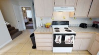 This two-story home offers a practical and straightforward layout designed for everyday living. The kitchen serves as a central feature, showcasing light wood cabinetry that extends to the upper walls, providing ample storage throughout. Tile countertops add a classic touch, while the electric coil range with an overhead ventilation hood anchors the cooking area. A small kitchen island with matching light wood cabinetry offers additional prep space and storage. The flooring throughout is light-toned tile, which contributes to a bright and cohesive feel. Lemon-themed decorative accents on the backsplash add a subtle charm to the cooking space. An open passageway from the kitchen provides a direct sightline into an adjacent bedroom, suggesting a connected and accessible floor plan. Natural light filters through the home, visible in the background rooms, enhancing the overall sense of openness. The ceiling fan glimpsed in the adjoining room indicates comfortable climate management throughout. Overall, the home presents a functional, no-frills interior well-suited for practical daily use.