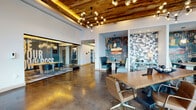 This single-floor coworking space delivers a thoughtfully designed environment that blends industrial aesthetics with contemporary comfort. The main open area is anchored by polished concrete floors and soaring ceilings clad in warm reclaimed wood planks with integrated ambient uplighting. A dramatic multi-bulb chandelier hangs at the center, casting a welcoming glow over a large live-edge communal table surrounded by mid-century modern chairs. Floor-to-ceiling glass walls and entry doors flood the space with abundant natural light and offer unobstructed street views, reinforcing a strong connection to the surrounding urban environment. Distinctive typographic murals in teal, gold, and neutral tones serve as bold focal points throughout the lounge, paired with hexagonal tile accent walls that add visual texture. Tufted teal booth seating provides semi-private nooks for focused work or informal meetings, each backed by typographic wallcoverings and equipped with wall-mounted displays. A glass-enclosed conference room offers a dedicated private meeting area furnished with task chairs and professional audiovisual equipment. A separate enclosed conference room features a motivational wall mural, a mounted flat-screen TV, a textured 3D tile accent wall, and stylish pendant lighting, creating an inspiring setting for presentations and team collaboration. Throughout, curated artwork, lush potted plants, and layered lighting contribute to a dynamic yet inviting professional atmosphere.