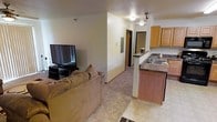 This single-floor space offers a functional and comfortable layout centered around an open-concept living and kitchen area. The living room is carpeted and well-appointed, featuring a ceiling fan with a light fixture for year-round comfort, a wall-mounted air conditioning unit, and large vertical-blind-covered sliding doors that allow natural light to filter into the room. The space accommodates generous seating with plush sofas arranged around a glass-top side table, creating a relaxed and inviting atmosphere.

The kitchen is positioned adjacent to the living area and is defined by a raised breakfast bar with a dark countertop that serves as a natural divider between the two spaces. Warm honey-toned wood cabinetry lines the upper walls, complemented by a built-in sink. Tile flooring in the kitchen transitions seamlessly to carpet in the living area, adding a practical distinction between zones.

A hallway visible from the living space leads toward additional rooms, suggesting a well-connected floor plan. Recessed lighting in the transitional area provides consistent illumination throughout. Overall, the space presents a straightforward, functional design suited for everyday living.