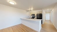This thoughtfully laid-out single-floor residence offers three bedrooms, two bathrooms, and a functional open-concept living space. The kitchen and living room flow seamlessly together, anchored by a peninsula-style counter with an integrated sink — ideal for both cooking and casual dining. The kitchen is equipped with black appliances, light wood cabinetry, and track lighting, all set atop warm light hardwood flooring that extends throughout the main living areas. A dedicated entrance leads into the space, which maintains a clean, bright, and neutral palette throughout.

The bedrooms are carpeted for comfort, with each room featuring its own closet. One bedroom includes a walk-in closet fitted with wire shelving for organized storage. The primary bedroom offers a wall-mounted mini-split heating and cooling unit along with direct access to a private outdoor area through a sliding glass door, bringing in natural light and outdoor views. Additional bedrooms also feature wall-mounted climate control units and windows with vertical blinds.

The bathrooms are finished with light hardwood-style flooring, clean white walls, and a tub-shower combination. The overall design emphasizes a bright, minimalist aesthetic with consistent finishes, making the space feel cohesive and move-in ready.