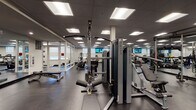 This fitness center offers a comprehensive, professionally designed workout facility spread across a single floor. The space is organized into distinct functional zones, each catering to different training needs. The main floor is finished with durable dark rubber flooring throughout, providing a clean and practical surface for all activities.

The cardio zone features a row of treadmills and elliptical trainers positioned along a windowed wall, with a wall-mounted television overhead for entertainment during workouts. Adjacent to this area, a selection of stationary cycling bikes occupies its own open section of the floor.

The strength training area is well-appointed with a full lineup of Matrix cable and resistance machines, along with a comprehensive dumbbell rack displaying a wide range of weights, paired with adjustable benches. Mirrored walls expand the visual depth of the space and support proper form during exercise.

A glass-walled enclosed studio room provides a dedicated space for flexibility training, yoga, or small group workouts, outfitted with stability balls and additional accessories. Vibrant botanical artwork decorates the teal-accented glass partition, adding a pop of color to the otherwise neutral palette.

The facility is brightly illuminated by recessed panel lighting set within a drop ceiling, creating an evenly lit environment. Entry is through a set of glass double doors, with clear sightlines across the entire floor plan ensuring an open and accessible atmosphere throughout.