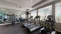 Located at 200 Village Blvd in Davenport, Florida, this well-equipped fitness center offers a comprehensive workout environment on a single floor. The space is thoughtfully designed with tray ceilings accented by crown molding and ceiling fans, lending the room a polished, comfortable atmosphere. Large windows with horizontal blinds line the walls, flooding the space with natural light and providing views of the surrounding outdoor landscape.

The cardio zone features multiple treadmills, elliptical trainers, and stationary bikes arranged along the windowed walls, creating an airy environment for aerobic exercise. Full-length mirrors are strategically positioned throughout the space, visually expanding the room and allowing users to monitor their form during workouts.

The strength training area is well-stocked with a rack of free weights spanning a range of sizes, accompanied by benches for targeted exercises. A multi-station cable machine supports a variety of resistance exercises, while a dedicated functional training zone — marked as a "Training Zone" — includes a wall-mounted training rig with TRX suspension equipment and medicine balls for dynamic, full-body workouts.

The space is finished with durable, dark carpet flooring throughout and is accessible via glass-paned French doors as well as an exterior door. A wall-mounted television is also present, adding to the overall experience of the facility.