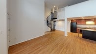 This two-floor loft-style space offers a thoughtfully designed layout with distinct character across both levels. The main floor features an open living area with warm hardwood-style flooring and soaring double-height ceilings that create an airy, expansive atmosphere. A dramatic black spiral staircase serves as both a functional and architectural centerpiece, connecting the two floors with visual flair. Large windows fitted with horizontal blinds flood the main living area with natural light. The kitchen is well-equipped with rich wood cabinetry, black appliances including a range, microwave, and dishwasher, complemented by recessed lighting and additional counter seating. The bathroom on the main level features light tile flooring, a long white vanity with an integrated sink, a large mirror, and a walk-in shower with built-in shelving. The upper level offers a carpeted bedroom retreat with impressively tall windows that draw in generous daylight. A secondary lower-level room provides additional carpeted space with clerestory windows, suitable for a variety of uses. A dedicated closet area with wire shelving and an adjacent vanity sink offers practical storage and grooming convenience. Throughout, the space maintains a clean, neutral palette that is both modern and versatile.