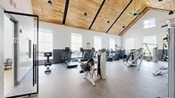 Valley View Gym in East Hanover, NJ is a thoughtfully designed fitness facility that combines modern architecture with a comprehensive range of exercise equipment. The main workout floor is the centerpiece of the space, featuring soaring vaulted ceilings clad in warm wood paneling that create an open, energizing atmosphere. Rows of large windows along the perimeter flood the floor with natural light while offering exterior views, and pendant lights and ceiling fans complement the airy aesthetic. Durable speckled rubber flooring runs throughout the main area, which is equipped with treadmills, ellipticals, stair climbers, and a variety of strength machines. Full-length mirrors along one wall enhance the sense of space and allow users to monitor their form. A dedicated cycling studio adjoins the main floor, featuring spin bikes on hardwood flooring, a large frosted window for natural light, and a pass-through window connecting it to the main gym. A separate strength and free weights area is accessible through a sleek glass pivot door and features textured wall finishes, additional benches and equipment, and interior windows that maintain visual connectivity throughout the facility. The overall design balances functionality with a clean, contemporary aesthetic suited for a serious fitness environment.