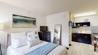 Located at 13223 Champions Centre Dr in Houston, TX, this efficiently designed studio offers a well-organized single-floor layout that maximizes every square foot. The open-concept floor plan seamlessly integrates the sleeping, living, and kitchen areas into one cohesive space, making it both functional and practical.

The bedroom area is generously proportioned for the overall footprint, accommodating a full-size bed with ample room to move around. A textured ceiling adds subtle architectural interest, while a window with blinds allows natural light to brighten the space. Decorative wall art and a full-length leaning mirror contribute to the room's clean, contemporary aesthetic. Light wood-style flooring runs throughout, lending warmth and visual continuity.

The kitchenette is thoughtfully appointed with dark espresso cabinetry, white countertops, a stainless steel sink, and overhead cabinets for storage. A range hood, microwave, and open shelving complete the compact but capable cooking space.

The bathroom is clean and straightforward, featuring a shower-tub combination with a curtain rod, toilet, and a wall-mounted vanity with a light-colored countertop. Light flooring continues from the main area, maintaining a cohesive design throughout.

The overall ambiance is bright, modern, and well-maintained, making this studio a practical and comfortable option in the Champions area of Houston.