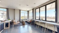 Located at 2501 N Orange Blossom Trail in Orlando, FL, this contemporary amenity lounge offers a thoughtfully designed communal space spread across a single floor. Large, floor-to-ceiling windows with dark metal frames flood the interior with abundant natural light while framing sweeping panoramic views of the Orlando skyline and surrounding landscape. The polished concrete floors give the space a sleek, modern industrial character that ties the interior together seamlessly.

The main lounge area features multiple distinct seating arrangements, including a casual conversation grouping with upholstered chairs, a central coffee table, and a built-in counter-height bar along the window wall, complete with woven bar stools ideal for working or socializing. A secondary lounge area is anchored by a wood-accented feature wall, a mounted flat-screen television, and comfortable lounge chairs arranged for relaxation or informal gatherings. A wine cooler and serving counter add functional convenience.

The hallway entrance is enlivened by a large, vibrant abstract artwork, creating a striking first impression. A restroom is conveniently accessible from this corridor. The space extends outdoors to a spacious terrace adorned with string lights, a dining table, and additional bar seating — all oriented toward the open sky and city views below, making it an ideal setting for both casual and social gatherings.