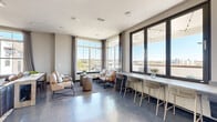 Located at 2501 N Orange Blossom Trail in Orlando, FL, this contemporary amenity lounge offers a thoughtfully designed communal space spread across a single floor. Large, floor-to-ceiling windows with dark metal frames flood the interior with abundant natural light while framing sweeping panoramic views of the Orlando skyline and surrounding landscape. The polished concrete floors give the space a sleek, modern industrial character that ties the interior together seamlessly.

The main lounge area features multiple distinct seating arrangements, including a casual conversation grouping with upholstered chairs, a central coffee table, and a built-in counter-height bar along the window wall, complete with woven bar stools ideal for working or socializing. A secondary lounge area is anchored by a wood-accented feature wall, a mounted flat-screen television, and comfortable lounge chairs arranged for relaxation or informal gatherings. A wine cooler and serving counter add functional convenience.

The hallway entrance is enlivened by a large, vibrant abstract artwork, creating a striking first impression. A restroom is conveniently accessible from this corridor. The space extends outdoors to a spacious terrace adorned with string lights, a dining table, and additional bar seating — all oriented toward the open sky and city views below, making it an ideal setting for both casual and social gatherings.