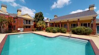 Located at 10027 Spice Ln in Houston, Texas, this residential community showcases traditional brick architecture with distinctive yellow accent trim, columned facades, and pitched rooftops with chimneys — elements that lend the property a classic, Southern-inspired aesthetic. The multi-building layout is set across a single floor plan and is connected by wide concrete walkways that guide residents through thoughtfully landscaped grounds.

At the heart of the community is a generously sized outdoor swimming pool with striking turquoise water, framed by a brick-trimmed pool deck and iron fencing. The pool area is complemented by a covered pavilion structure with shade sails overhead, offering a comfortable outdoor retreat. Surrounding the pool, mature magnolia and palm trees, crepe myrtles, and manicured shrubs create a lush, inviting atmosphere that thrives under the Texas sun.

The common outdoor areas feature well-maintained pathways bordered by natural landscaping, including decorative rock arrangements and garden beds. Iron fencing delineates key areas of the property, adding both structure and charm. The buildings feature screened balconies and large windows that enhance natural light and ventilation. Overall, the community presents a cohesive, well-kept environment ideal for comfortable residential living in the Houston area.