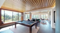 This single-level clubhouse offers a thoughtfully designed social hub with a variety of distinct activity and relaxation zones. The space is anchored by a striking wood-slatted ceiling that runs throughout, adding warmth and architectural continuity. Floor-to-ceiling windows and curved bay window nooks flood the interiors with natural light and frame verdant, tree-lined views from multiple vantage points.

The game room serves as a central focal point, featuring a full-size billiards table set beneath industrial-style pendant lights, with a dedicated poker table nearby enclosed by a dramatic curved sheer curtain partition that rises to an open upper-level mezzanine. Decorative geometric screen panels and vertical wood slat dividers elegantly separate the zones while maintaining an open, airy flow throughout.

A cozy lounge area is furnished with tufted sofas, accent chairs in rich jewel tones, and a gold circular coffee table, creating an inviting gathering space. Built-in window benches with cushioned seating provide additional reading and relaxation spots. A casual bar and high-top seating area, along with a well-stocked shelving display, round out the amenities. Dark hardwood floors anchor the space, complementing the natural material palette. The overall ambiance is sophisticated yet welcoming, ideal for both social events and quiet leisure.