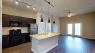 This 2-bedroom, 2-bathroom residence is laid out on a single floor, offering a functional and stylish living experience. The open-concept kitchen and living area serves as the heart of the home, featuring rich dark-espresso cabinetry, stainless steel appliances, and a classic white subway tile backsplash. A central kitchen island with a granite countertop and an undermount sink adds both prep space and a natural gathering point. Three sleek pendant lights hang above the island, providing focused task lighting with a contemporary flair, while track lighting enhances the kitchen further. Durable hardwood-style flooring runs throughout the main living area, adding warmth and cohesion. The adjoining living space is generously proportioned, bathed in natural light from a large window, and finished with a ceiling fan for year-round comfort. Neutral wall tones throughout create a bright and welcoming ambiance, offering a versatile backdrop for any décor style. The layout flows intuitively, with clear sightlines connecting the kitchen to the living area and access points leading to the bedrooms and bathrooms beyond.