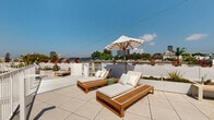 Located at 1503-1507 S Barrington Ave in the desirable West Los Angeles neighborhood of 90025, this rooftop deck offers an impressive open-air retreat with panoramic views of the surrounding cityscape. The space is finished with large-format light stone tiles that lend a clean, modern aesthetic throughout.

The rooftop is divided into distinct functional zones. A sun-drenched lounging area features teak chaise lounges with cushioned upholstery and a striped market umbrella, ideal for relaxing under the Southern California sky. Adjacent to this, a fire pit area with a glass-encased lava rock burner is surrounded by contemporary seating, creating an inviting gathering spot.

An outdoor kitchen and dining zone is equipped with built-in appliances and sleek white cabinetry, complemented by wood-accented bench seating. Lush planters with ornamental trees, palms, and colorful shrubs are integrated throughout, adding vibrant greenery against the bright white perimeter walls.

White metal railings line the perimeter, ensuring safety while preserving unobstructed sightlines to the Los Angeles skyline and neighboring architecture. The rooftop is accessible via a stairwell and benefits from abundant natural light throughout the day, making it a versatile and sophisticated outdoor amenity space.