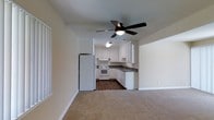 This single-floor residence offers a practical and well-organized layout designed for comfortable everyday living. The living area features neutral-toned carpeting and is equipped with a ceiling fan with integrated lighting, creating a relaxed and functional atmosphere. The space flows seamlessly into the kitchen, which is visually distinct with its rich dark hardwood-style flooring. The kitchen is outfitted with crisp white cabinetry, dark countertops, and white appliances — including a refrigerator, electric range, and dishwasher — all arranged in an efficient L-shaped configuration. Under-cabinet lighting adds a practical touch to the workspace. The kitchen also features a sink positioned along the cabinetry run, maximizing counter and storage space. Clean white walls and light-toned ceilings throughout the living and kitchen areas contribute to a bright, airy feel. The transition between the carpeted living space and the kitchen's hard flooring creates a clear and natural delineation between the two areas, while the open pass-through design keeps the overall layout feeling connected and spacious.