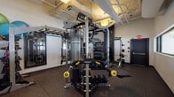 This single-floor fitness facility is thoughtfully designed to accommodate a wide range of training modalities. The main workout area is anchored by a professional-grade Hoist multi-functional cable and strength machine system, providing extensive options for resistance training. Complementing this centerpiece are free weights including kettlebells and barbells with weight plates, a dedicated weight bench, and organized dumbbell storage racks. A ceiling-mounted rig supports TRX suspension trainers and battle ropes, enabling functional and bodyweight training. Full-length mirrors mounted along one wall enhance spatial awareness during workouts and help visually expand the space. The flooring transitions between durable black rubber tiles in the primary training zones and striped carpet in transitional areas, offering both performance and comfort underfoot. Horizontal ribbon windows line one wall, allowing natural light to filter in and providing views into an adjacent well-lit space. A hydration station with an integrated water bottle filling system is conveniently located near the entrance. Stability balls and resistance bands round out the equipment offerings, making this a versatile and comprehensively outfitted fitness space.