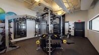 This single-floor fitness facility is thoughtfully designed to accommodate a wide range of training modalities. The main workout area is anchored by a professional-grade Hoist multi-functional cable and strength machine system, providing extensive options for resistance training. Complementing this centerpiece are free weights including kettlebells and barbells with weight plates, a dedicated weight bench, and organized dumbbell storage racks. A ceiling-mounted rig supports TRX suspension trainers and battle ropes, enabling functional and bodyweight training. Full-length mirrors mounted along one wall enhance spatial awareness during workouts and help visually expand the space. The flooring transitions between durable black rubber tiles in the primary training zones and striped carpet in transitional areas, offering both performance and comfort underfoot. Horizontal ribbon windows line one wall, allowing natural light to filter in and providing views into an adjacent well-lit space. A hydration station with an integrated water bottle filling system is conveniently located near the entrance. Stability balls and resistance bands round out the equipment offerings, making this a versatile and comprehensively outfitted fitness space.