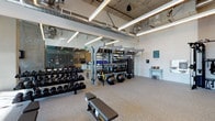 Located at 900 N La Brea Ave in Los Angeles, this contemporary fitness center combines industrial design with a well-equipped workout environment. The space features soaring ceilings with exposed concrete, structural beams, and visible ductwork, all illuminated by sleek linear LED lighting that creates a bright, energizing atmosphere.

The main workout floor is finished with durable rubber flooring and is organized into distinct functional zones. A dedicated TRX Training Zone anchors the space, housing a multi-tiered equipment rack stocked with medicine balls, stability balls, resistance tools, exercise mats, and TRX suspension trainers. Adjacent to this, a comprehensive free weight section features multiple rows of dumbbells alongside adjustable weight benches, catering to a full range of strength training needs.

The cardio zone is equipped with Life Fitness machines, including treadmills and upright bikes, positioned near floor-to-ceiling windows that invite ample natural light. A glass-walled reception and lounge area with warm lighting and comfortable seating provides a welcoming transition into the facility. Full-length mirrors along the walls enhance the sense of openness throughout the space. The overall design balances an urban, industrial aesthetic with a clean, functional layout suited for a comprehensive fitness experience.